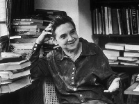 Adrienne Rich seated in front of a desk heaped with books, smiling.