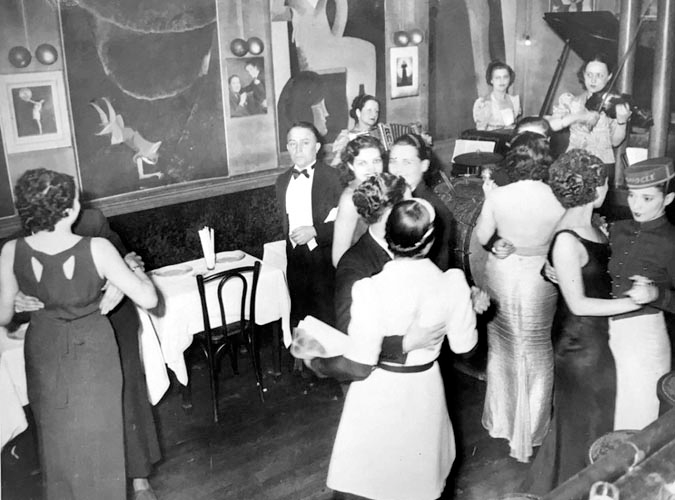 An intimate, black-and-white photograph of women dancing in one another's arms to a live band at a bar.